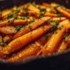 Bâtonnets de carottes dans une poêle en fonte, enrobés de beurre fondu, d'ail, et de miel, prêts à être enfournés.