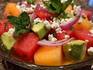 Ingrédients pour la salade estivale disposés sur une planche à découper : dés de melon, pastèque, avocat et oignon.
