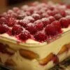 Une part de tiramisu framboise servie dans une assiette blanche, décorée d'une framboise fraîche et d'une feuille de menthe.