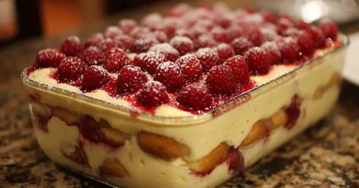 Une part de tiramisu framboise servie dans une assiette blanche, décorée d'une framboise fraîche et d'une feuille de menthe.
