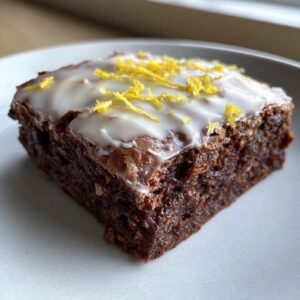 Gros plan d'une part de brownies au citron, glacée et garnie de zeste de citron.