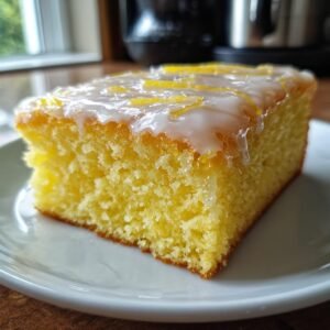 Gros plan d'une tranche de lemon brownies sur une assiette blanche, garnie d'un glaçage au citron et de zestes.